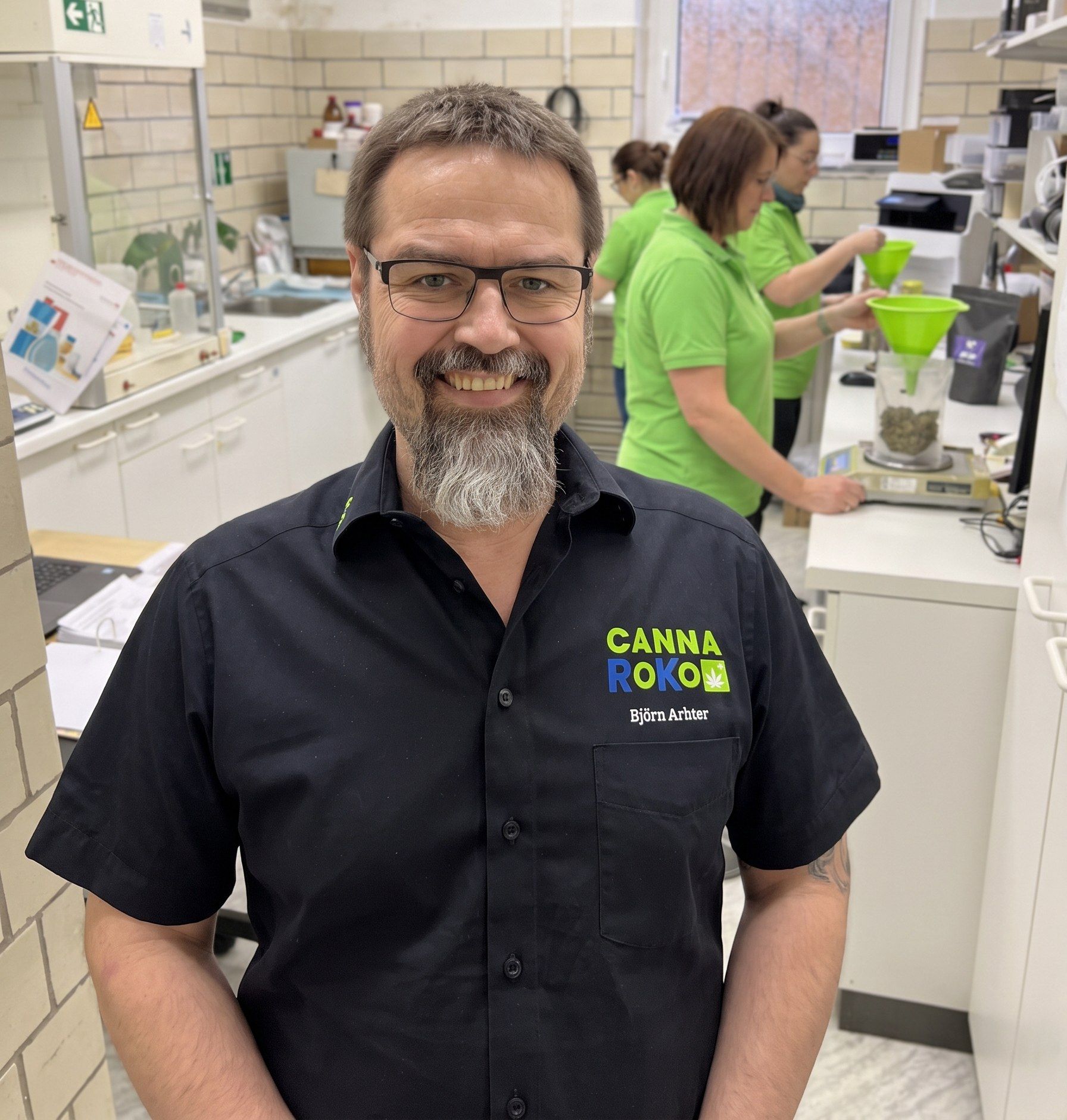 Apotheker Björn Arbter im CannaRoKo-Shirt, dahinter das Team beim Abwiegen der Medizinalcannabis-Blüten in der Robert-Koch-Apotheke