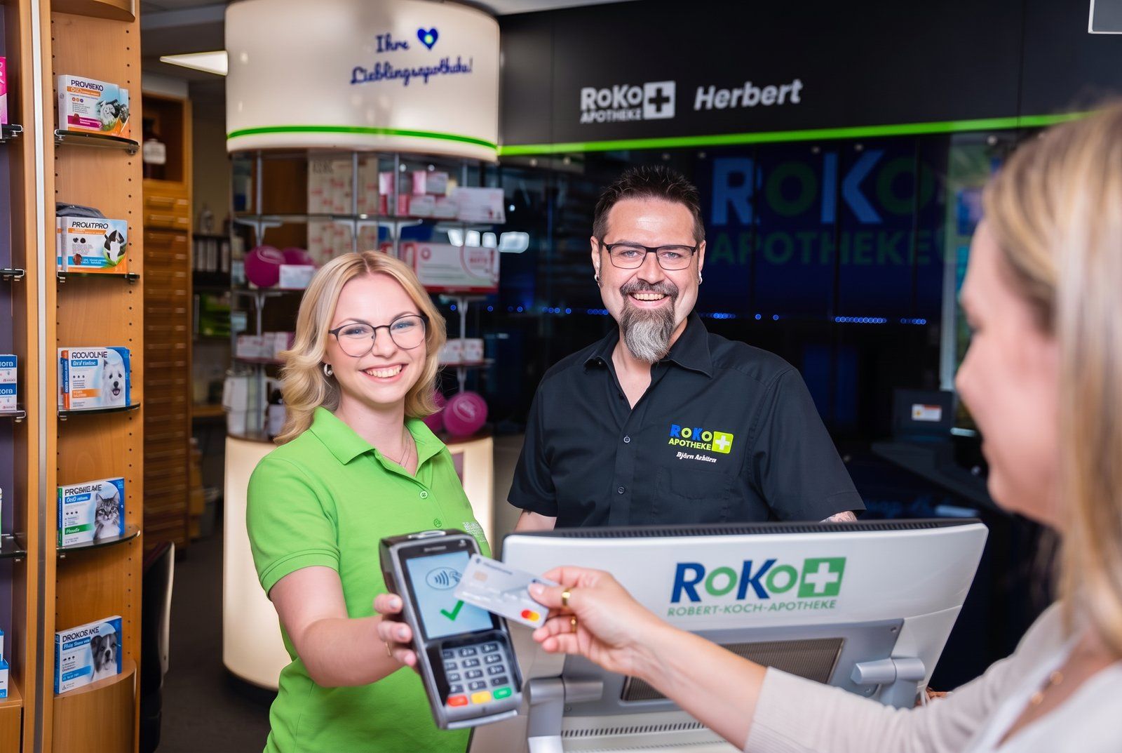 RoKo-Mitarbeiterin hält das Kartenterminal, eine Kundin bezahlt kontaktlos mit Karte an der Kasse der Robert-Koch-Apotheke